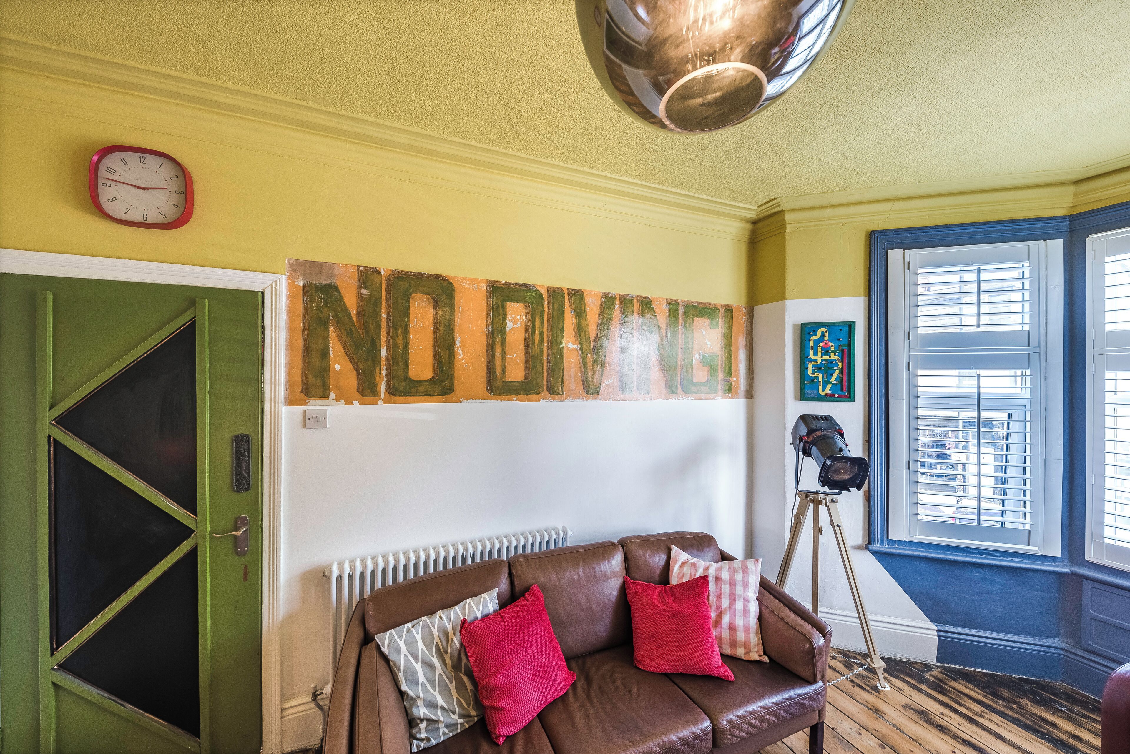 Une pièce avec un canapé en cuir marron, une porte verte, une horloge murale et un grand panneau indiquant « DÉBARQUEMENT INTERDIT ».