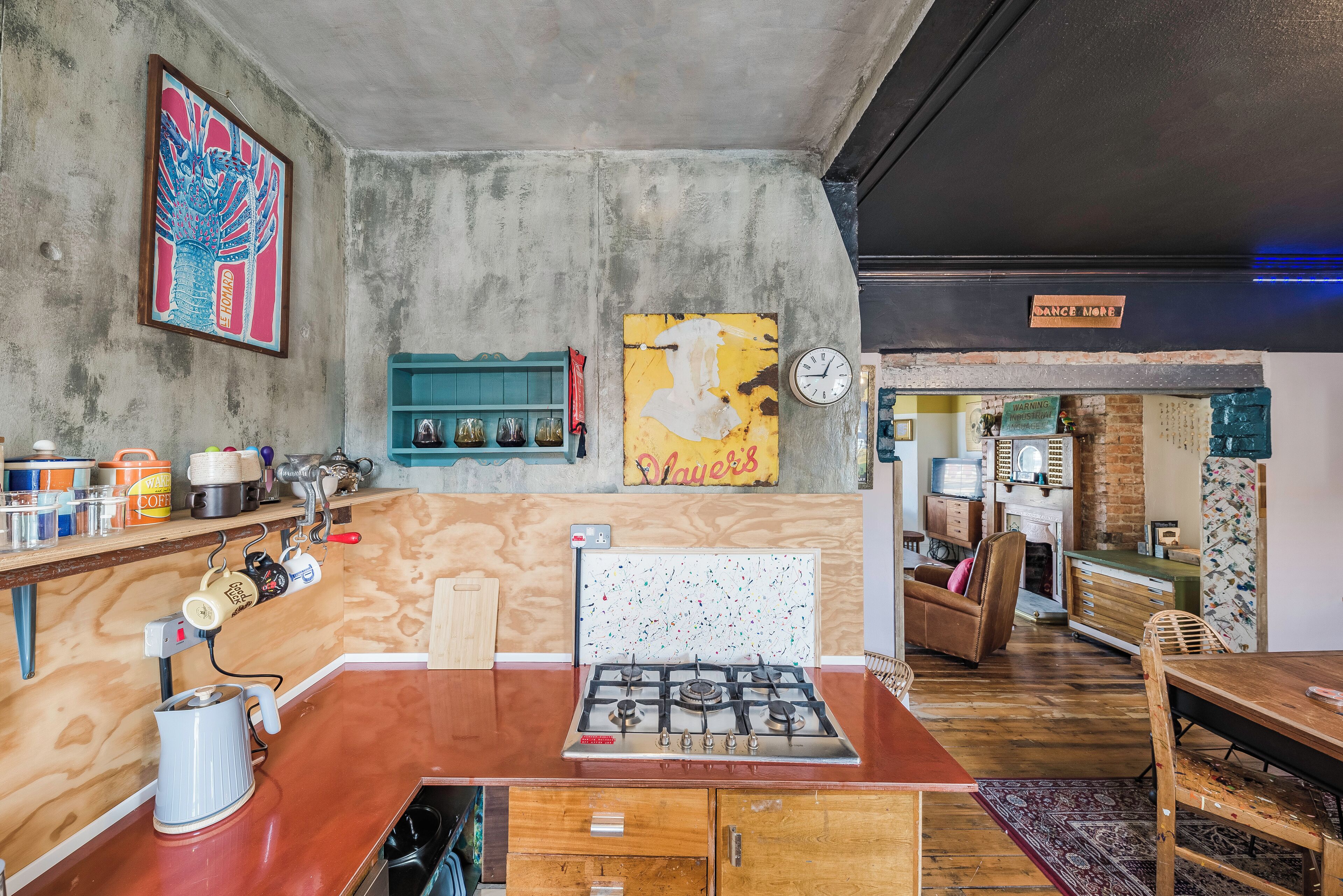 A kitchen with a gas stove, wooden cabinets, and a coffee machine. There is a clock on the wall and a sign that says "Dance More".