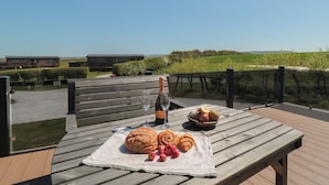 Une table en bois, sur laquelle sont posés une bouteille de champagne, un saladier de fruits et des pâtisseries, sur une terrasse offrant une vue sur les bâtiments et la verdure.