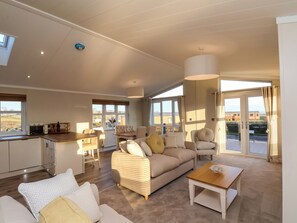 A spacious living room with a sofa, armchairs, a coffee table, and a dining area with a table and chairs. Large windows provide ample natural light and views of the outside. - Cherry Blossom Lodge (Whitby)