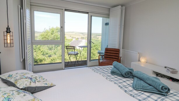 A bedroom with a large sliding glass door leading to a balcony, a bed with floral pillows, a blue checkered blanket, and a wooden chair. - Cysgod Y Garn (Newport)