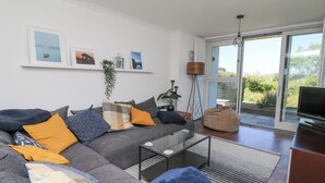 A modern living room with a grey sofa, blue and yellow cushions, a glass coffee table, and a television. There are framed pictures on the wall, a floor lamp, and a view through a sliding glass door. - Cysgod Y Garn (Newport)