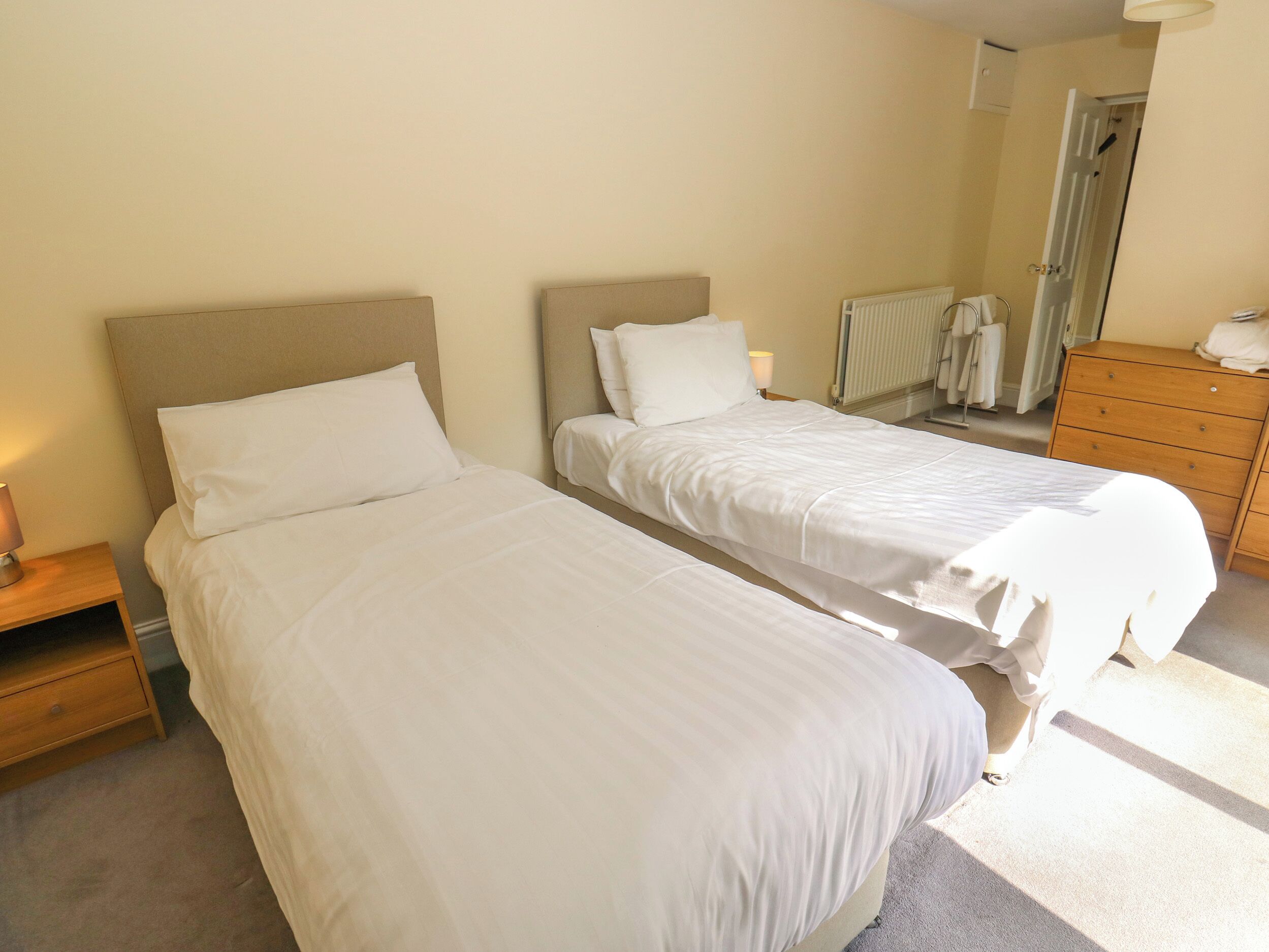 Une chambre d’hôtel avec deux lits, une commode en bois, un radiateur et une porte donnant sur une autre pièce.