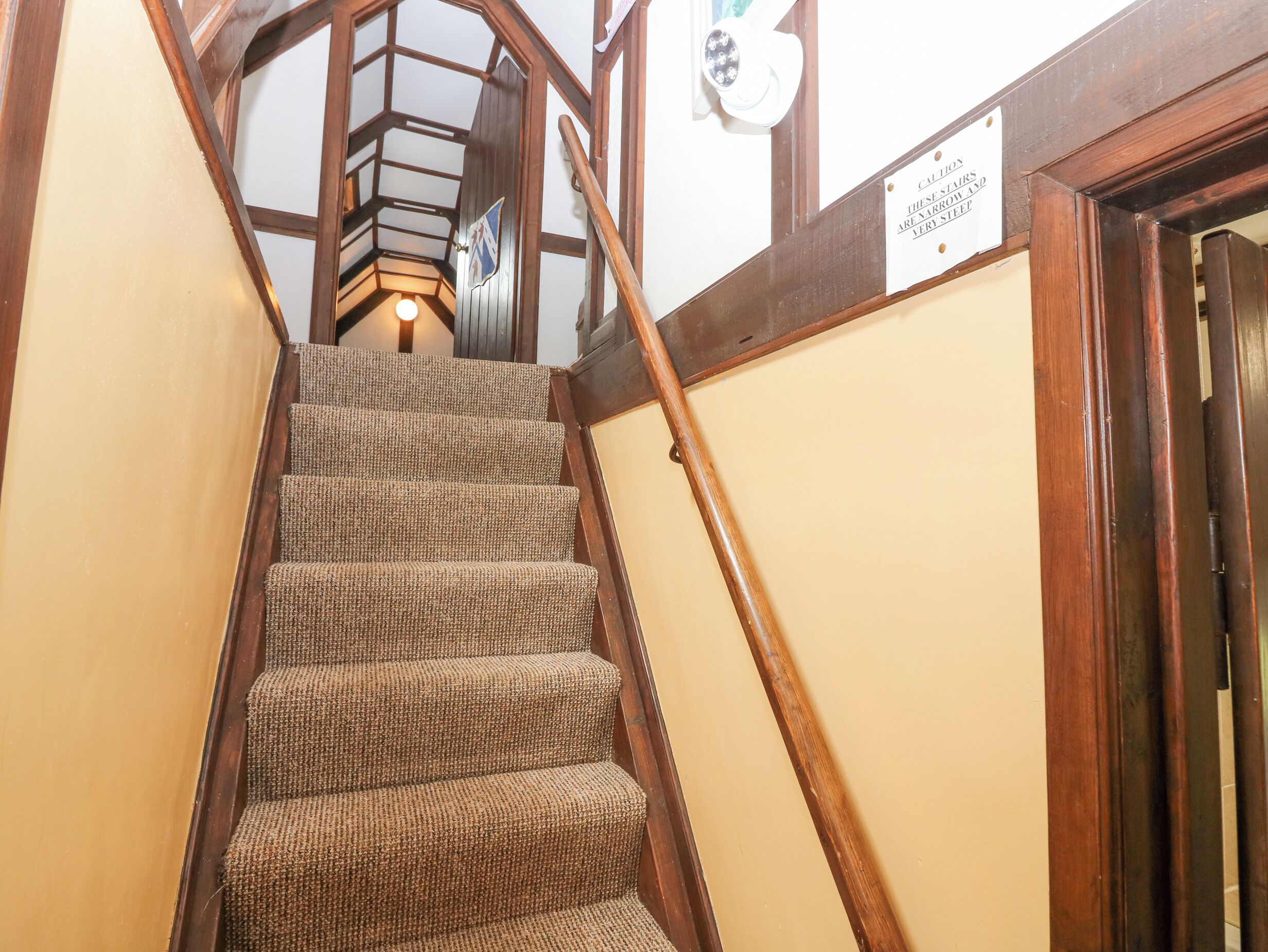 A wooden staircase with a handrail, carpeted steps, and a sign warning of narrow stairs.