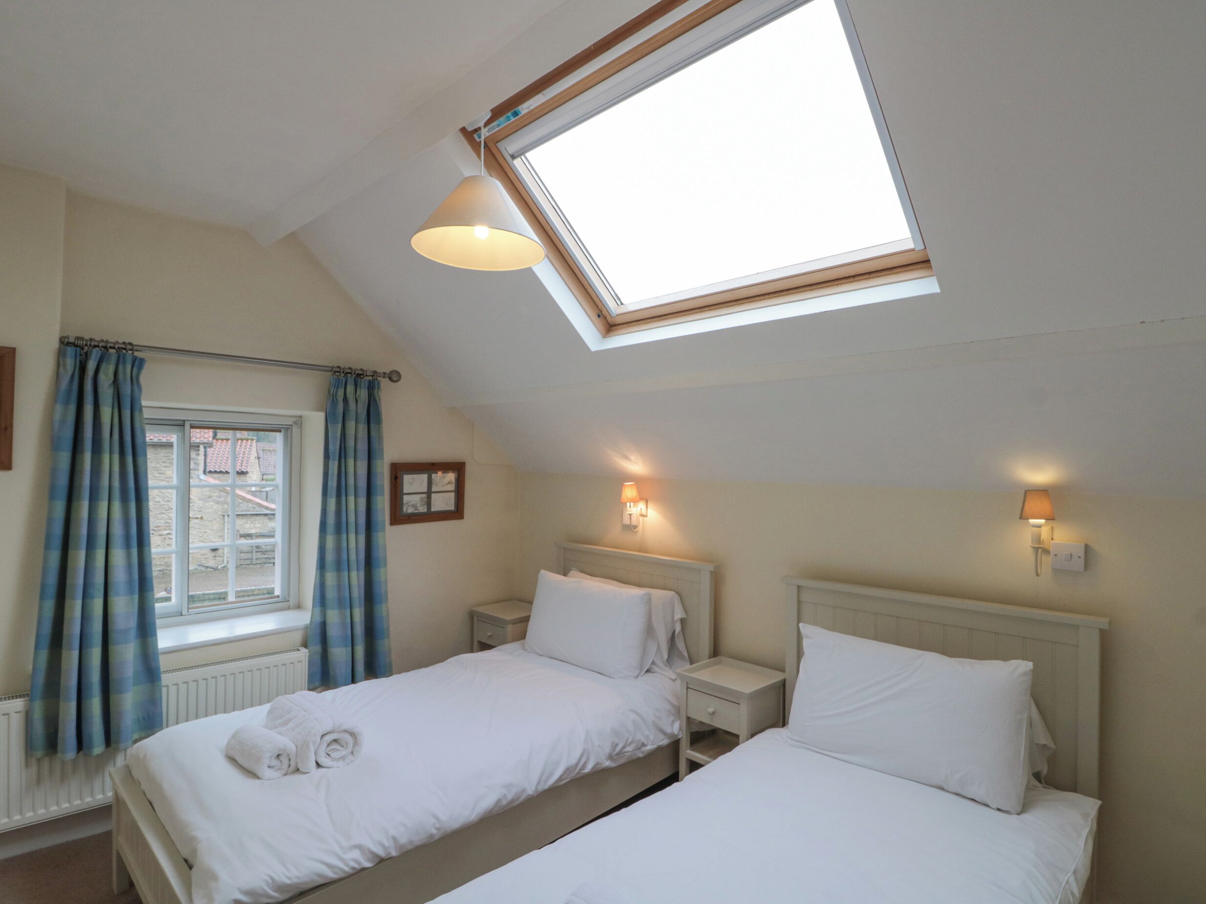 A bedroom with two beds, a skylight, a window with curtains, and bedside lamps.