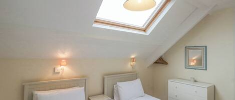 A bedroom with two beds, a skylight, bedside lamps, a dresser, and a mirror on the wall.