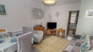Ein Wohnzimmer mit einem grauen Sofa, einem Esstisch gedeckt mit Tellern und Gläsern, einem Fernseher auf einem Holzschrank und einem gelben Teppich mit Muster.