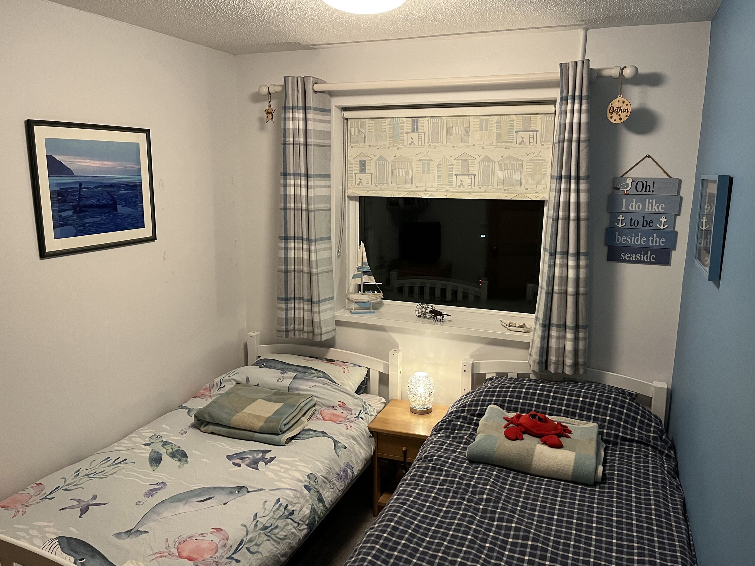 A bedroom with two beds, a window with curtains, a framed picture on the wall, and a small shelf with decorative items.
