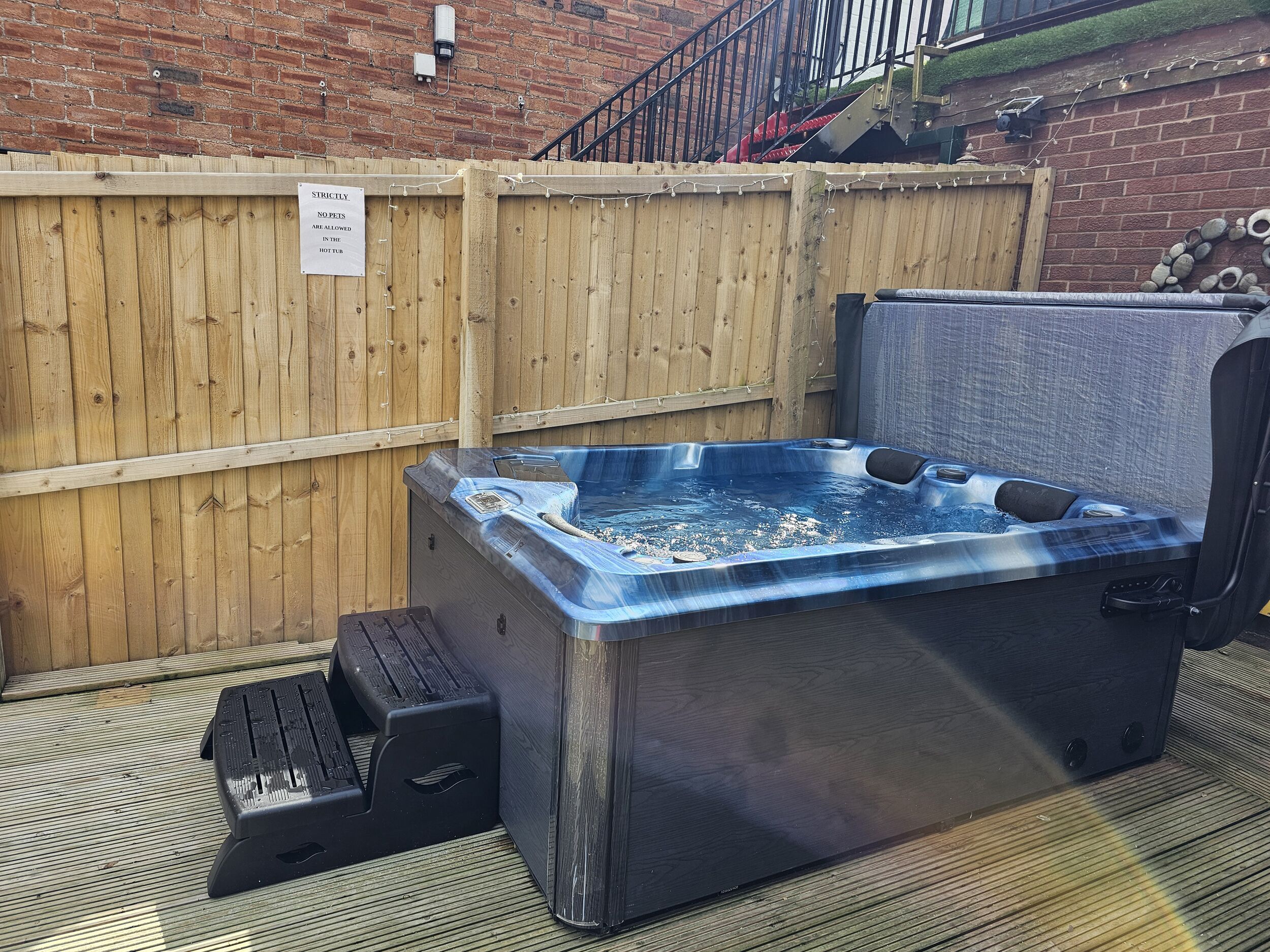 A hot tub with a wooden deck and a brick wall in the background.