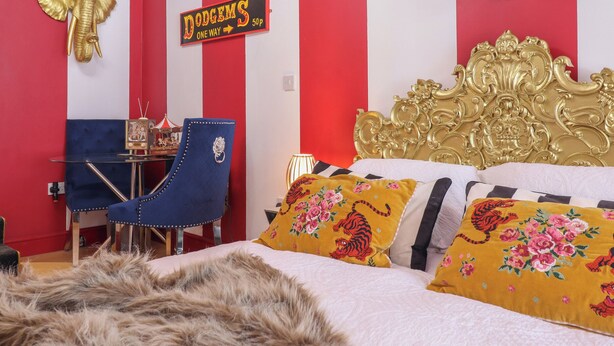 Une pièce avec un mur rayé rouge et blanc, un lit orné de coussins à motifs floraux, une chaise bleue, une décoration représentant un éléphant doré et une enseigne « Dodges ».