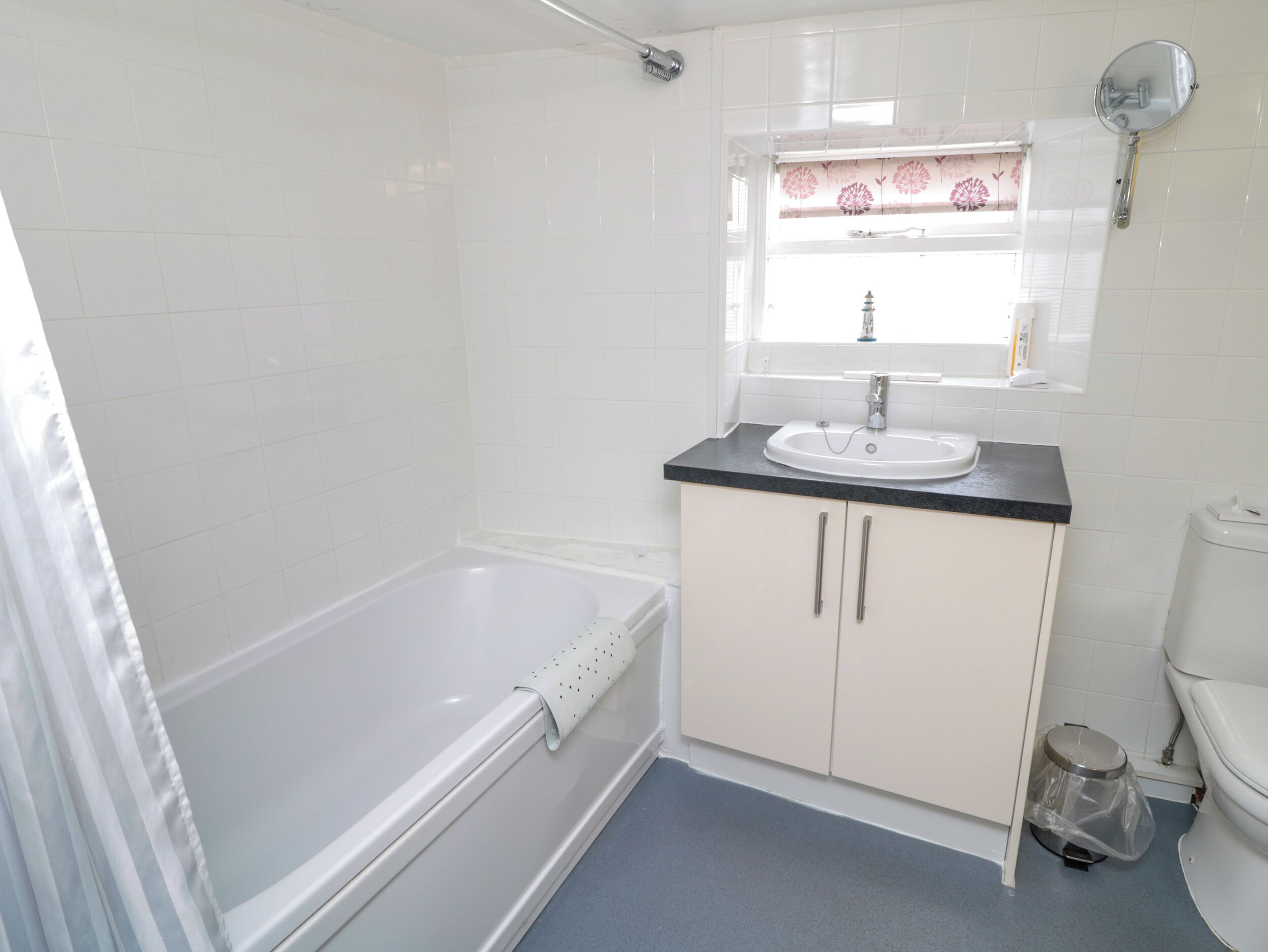 Une salle de bain avec une baignoire, un lavabo, des toilettes et une fenêtre ornée d’un rideau à motifs floraux.