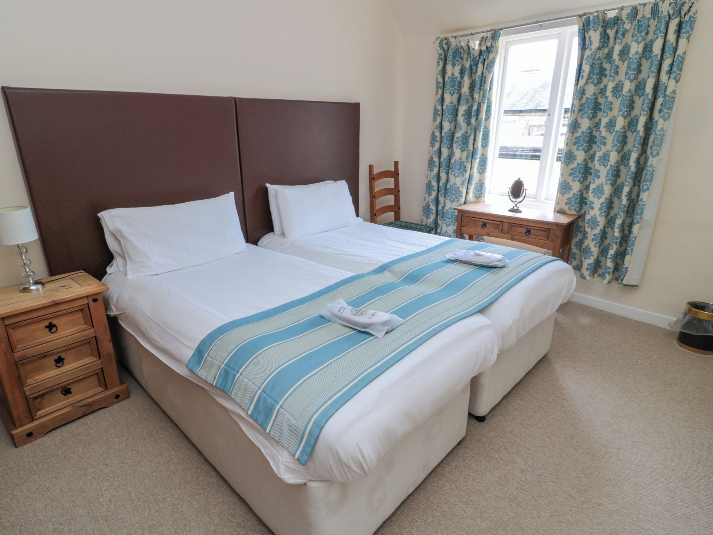 A double bed with a striped bedspread, wooden headboard, two bedside tables with lamps, a wooden chair, and a window with patterned curtains.