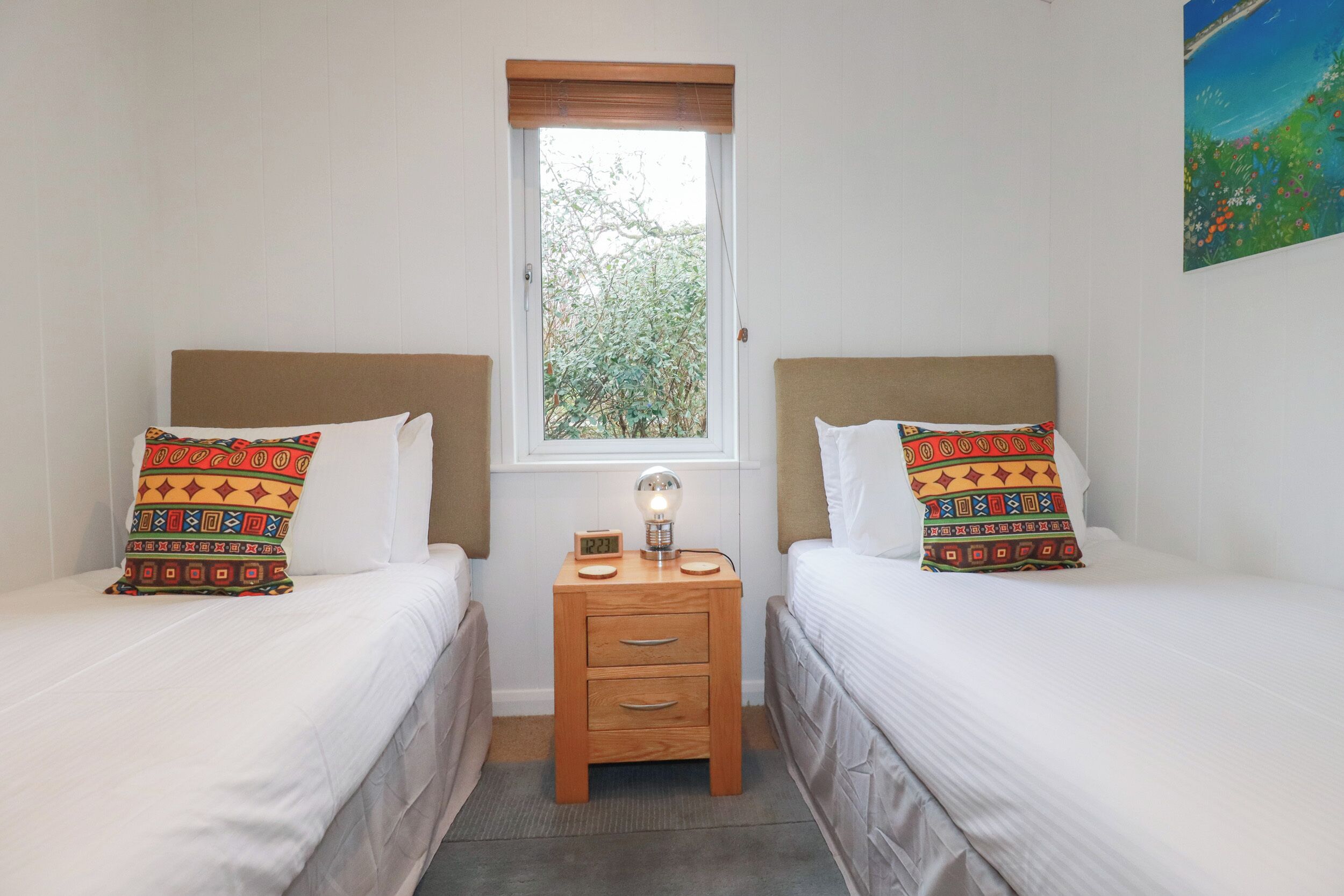 A bedroom with two beds, each with patterned pillows, a bedside table with a lamp, and a window with a view of greenery.