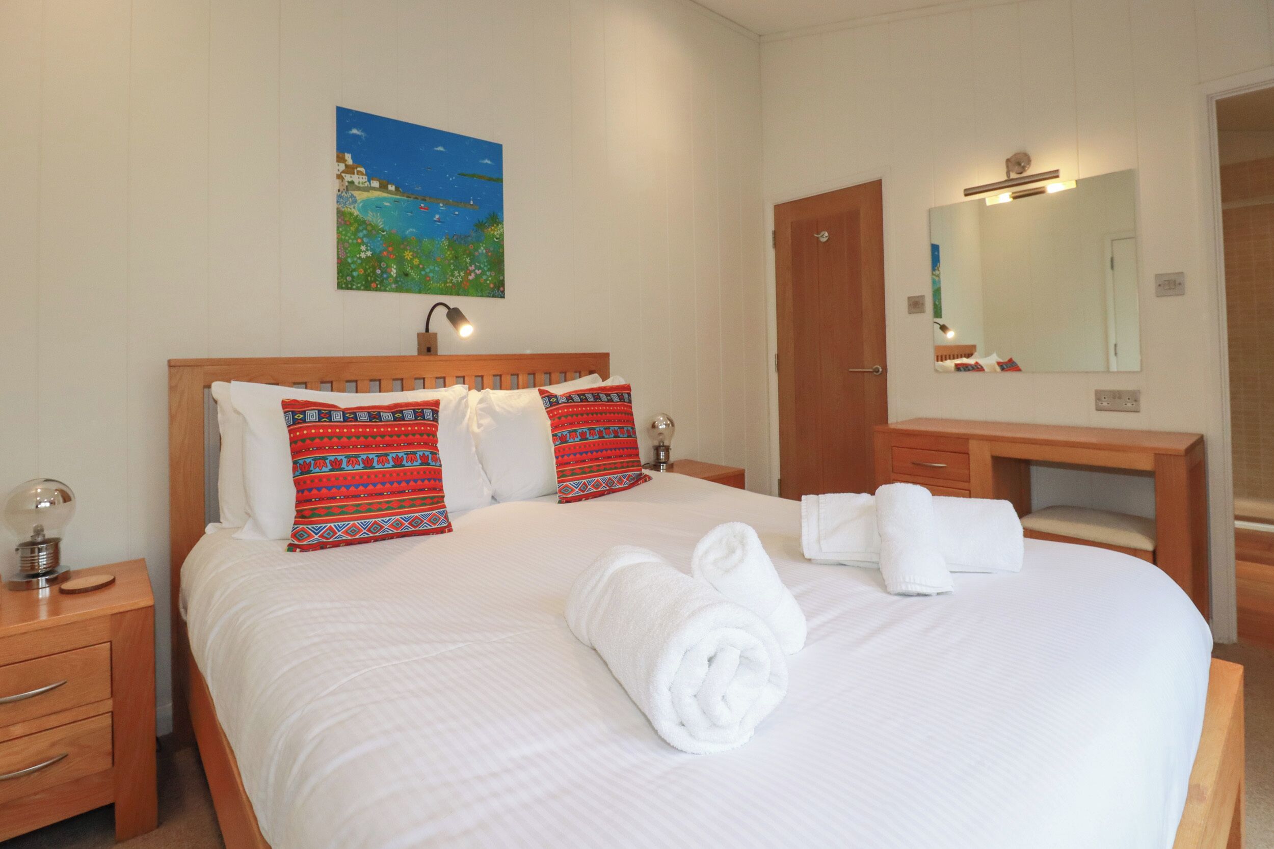 A neatly made bed with white linens and rolled towels, a wooden nightstand with a lamp, a wooden dresser, and a large mirror above the dresser.
