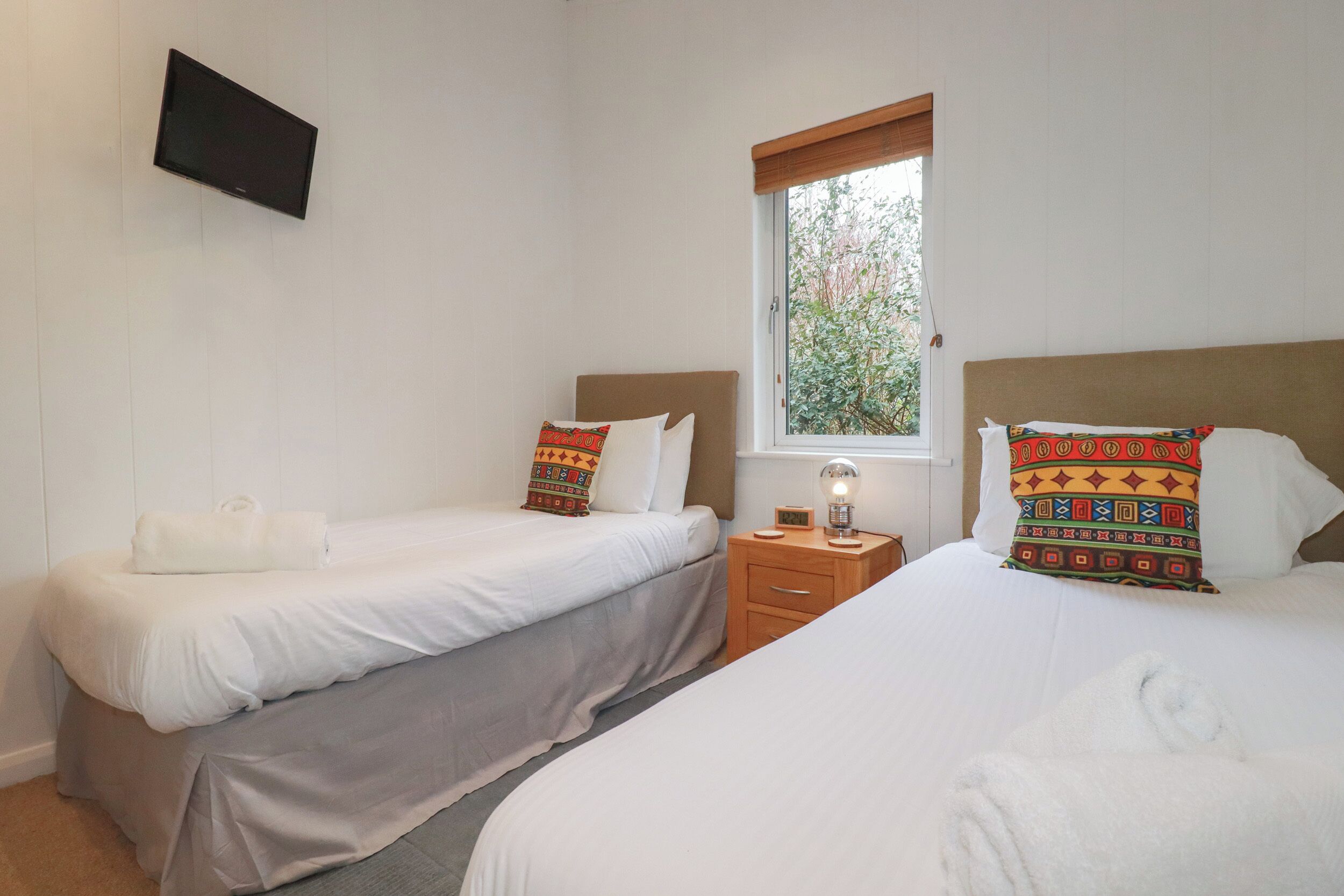 Une chambre d’hôtel avec deux lits simples, un téléviseur fixé au mur, une table de chevet avec une lampe et une fenêtre donnant sur un paysage verdoyant.