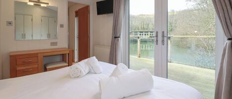A hotel room with a bed, a wooden dresser, a mirror, a television, and a balcony with a view of a body of water and trees.