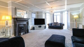 A spacious living room with a fireplace, a large flat-screen TV, a dark blue sofa, and a chandelier. - Maisonwyck (Canvey Island)