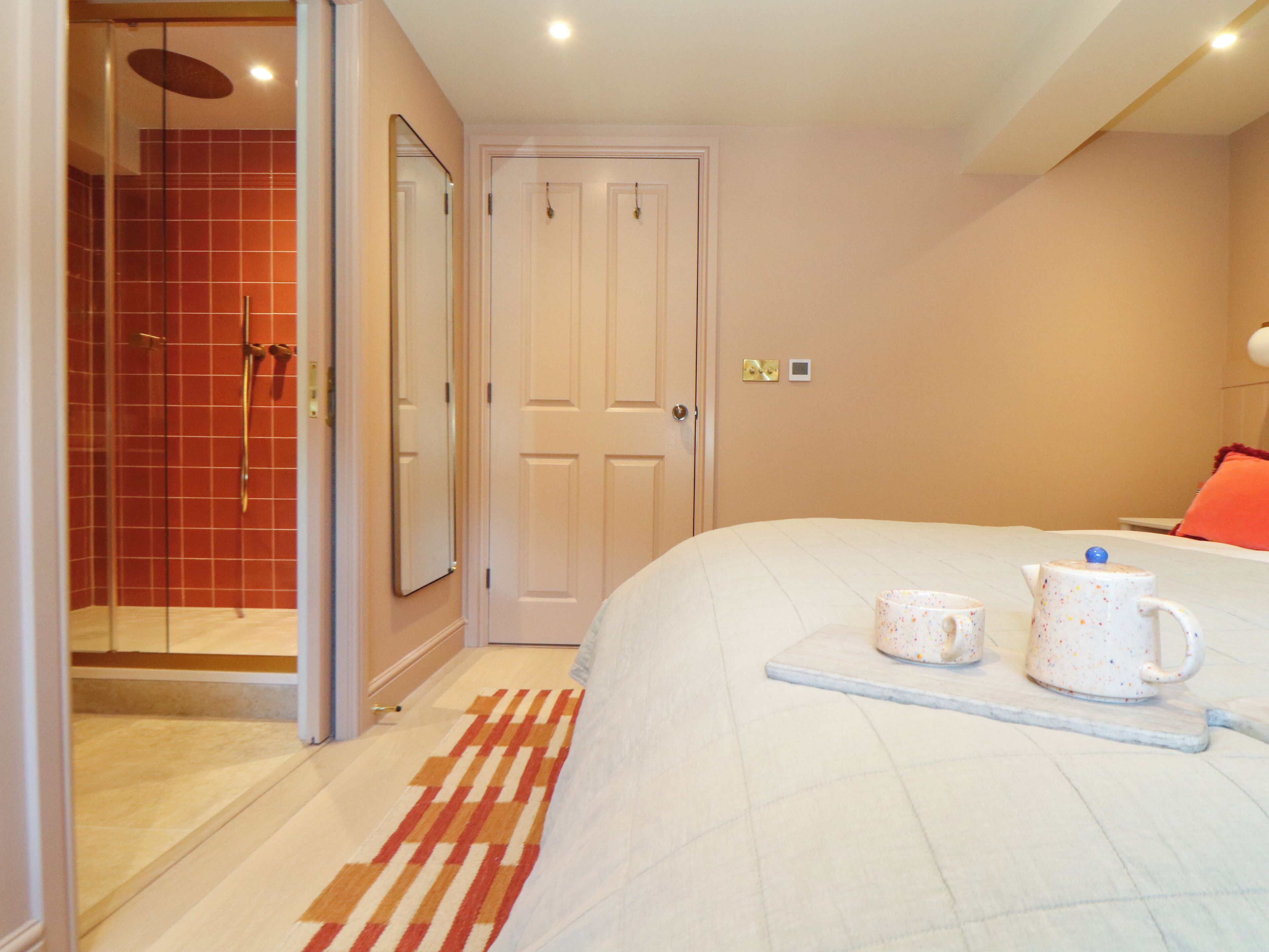 Une chambre à coucher avec un lit, une table de chevet sur laquelle sont posées deux tasses, un espace douche avec des carreaux rouges et une porte blanche.