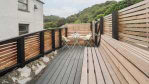 A wooden deck with a seating area, a white building, and a hillside in the background. - Mavericks Wave (St. Agnes)