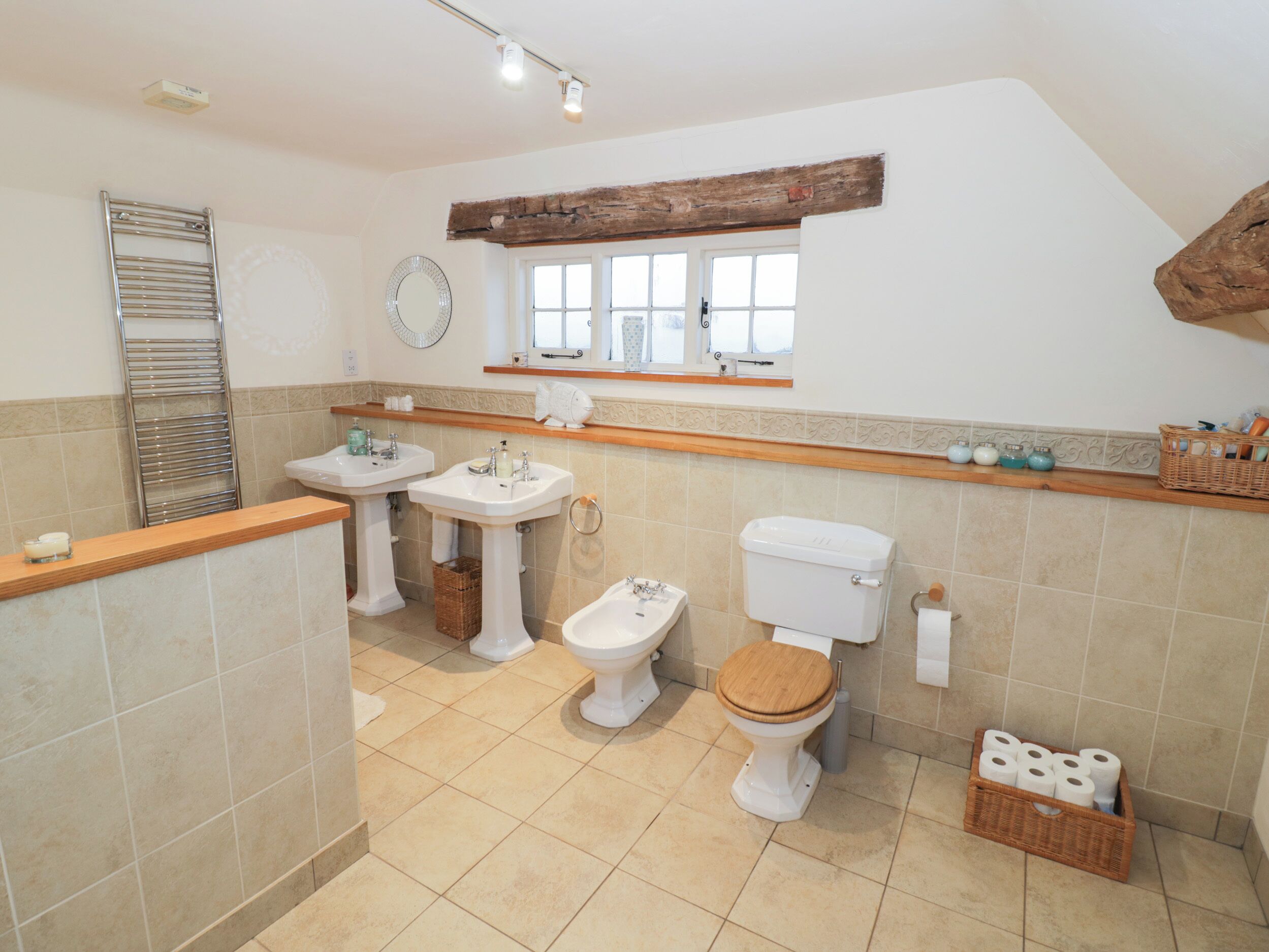 A bathroom with two sinks, a toilet, and a bidet. There is a wooden shelf above the sinks, a mirror, and a window.