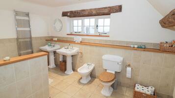 A bathroom with two sinks, a toilet, and a bidet. There is a wooden shelf above the sinks, a mirror, and a window.