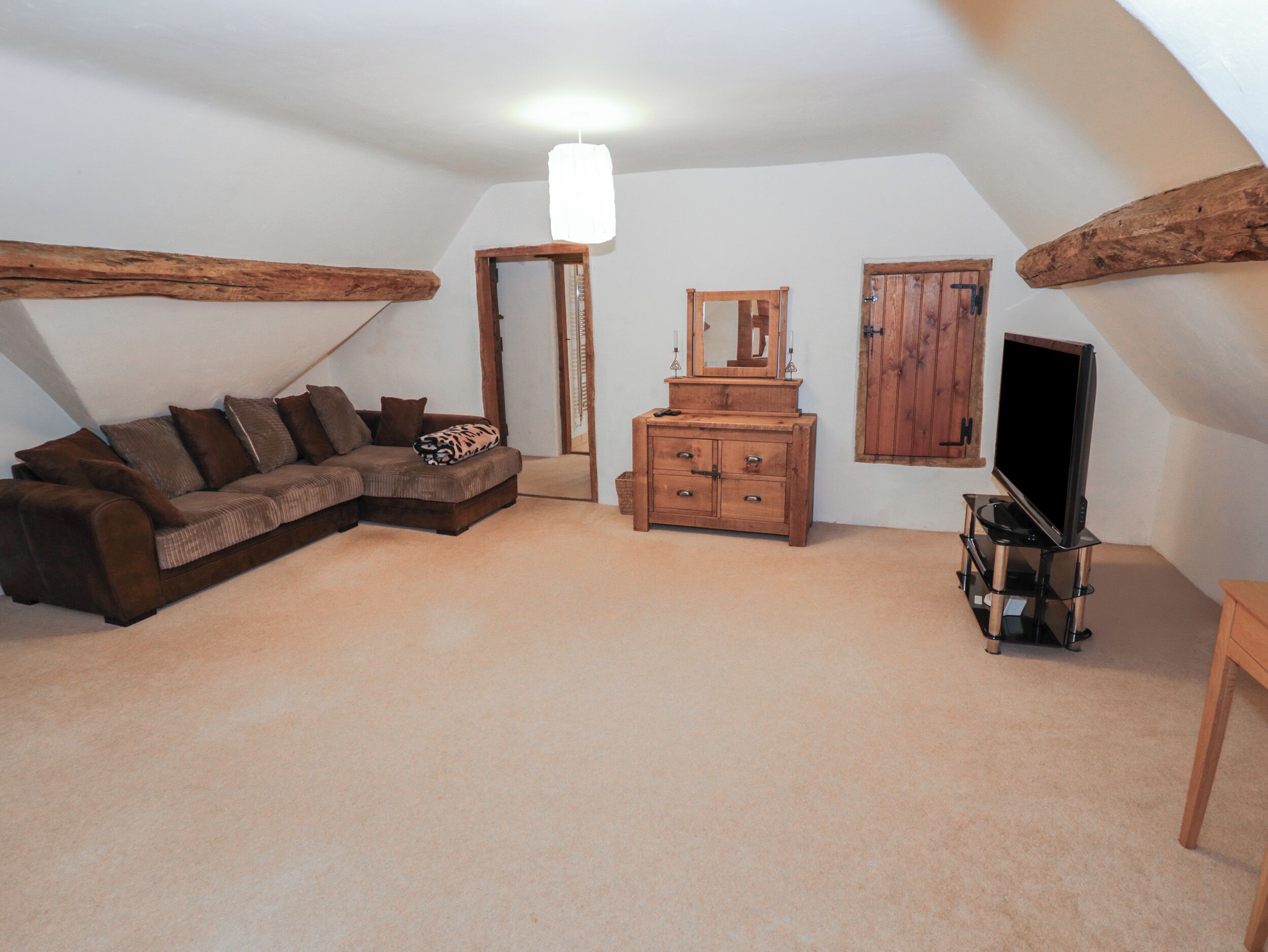 A cozy attic room with a brown sectional sofa, a wooden dresser, a television on a stand, and a large mirror.