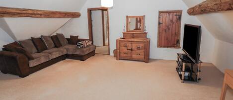 A cozy attic room with a brown sectional sofa, a wooden dresser, a television on a stand, and a large mirror.