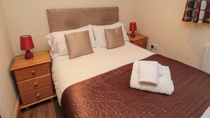 Una cama bien hecha con un edredón marrón, dos almohadas y una toalla doblada. Hay dos mesitas de noche con lámparas, una pared con tres cuadros enmarcados y una ventana con cortinas.