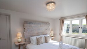 A bedroom with a wooden headboard, two bedside tables with lamps, a large window with curtains, and a radiator. - Beacon House (Whitby)