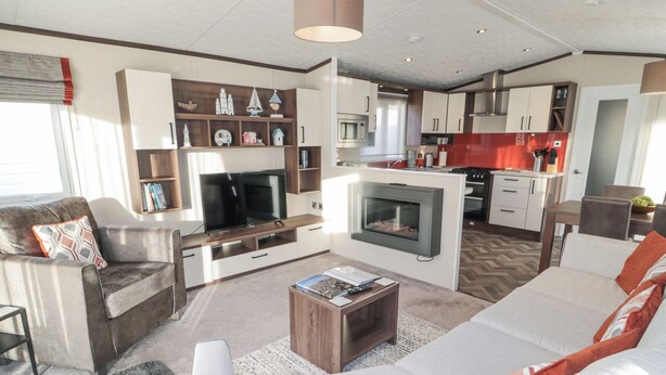 A modern living room with a sofa, coffee table, and entertainment unit. There is a kitchen area with cabinets and a microwave. - Downtime Lodge (Morpeth)
