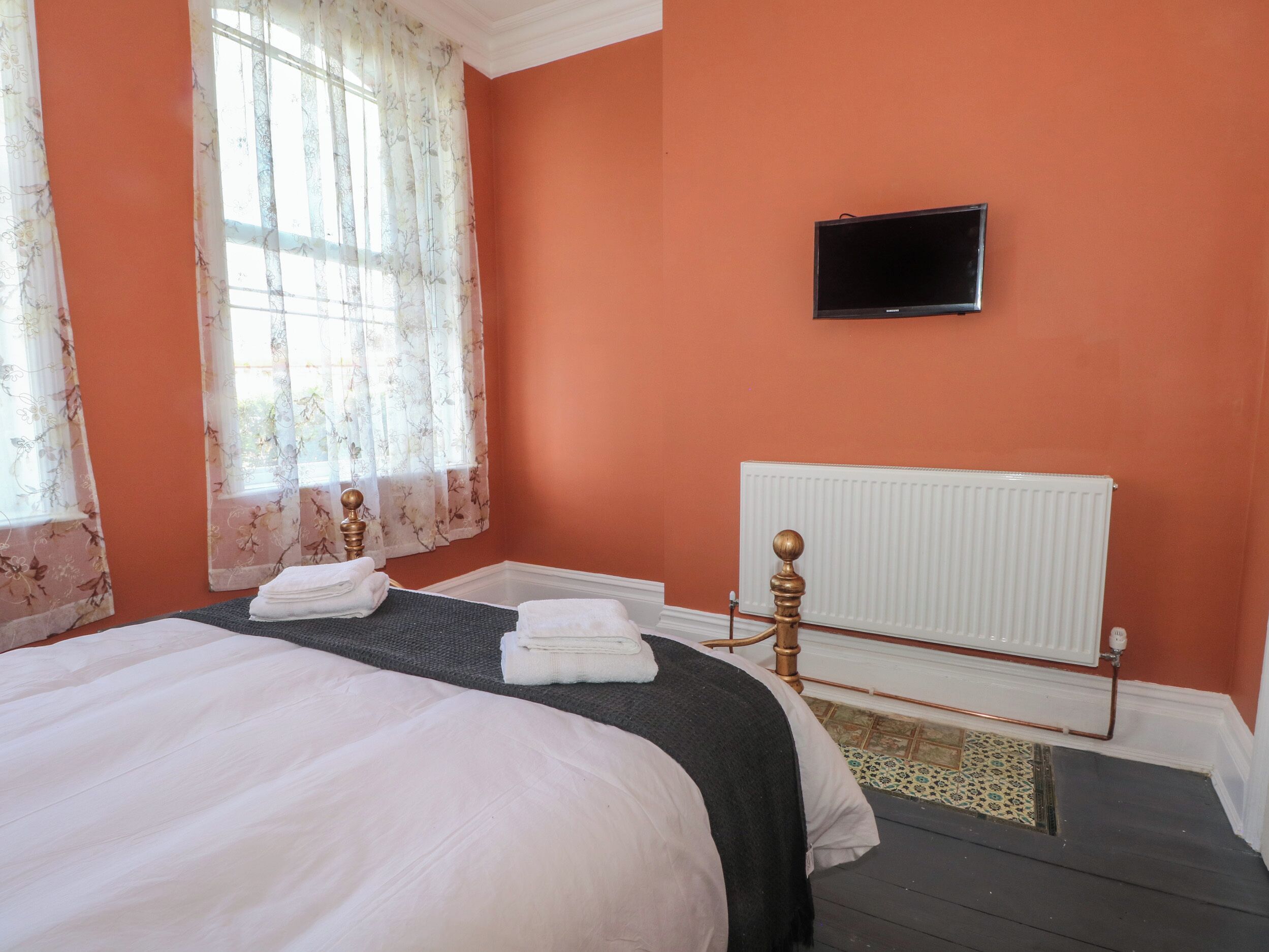 A bedroom with a bed, a radiator, a TV, and a window with floral curtains.