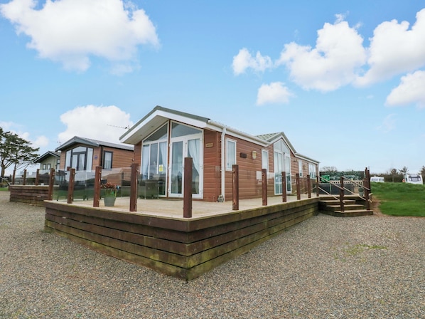 A wooden mobile home with a deck, large windows, and a gravel driveway.