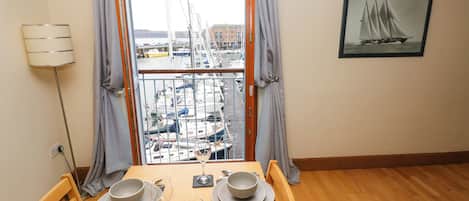 A dining area with a table set for two, a view of a marina with boats, and a framed picture of a sailing ship on the wall.