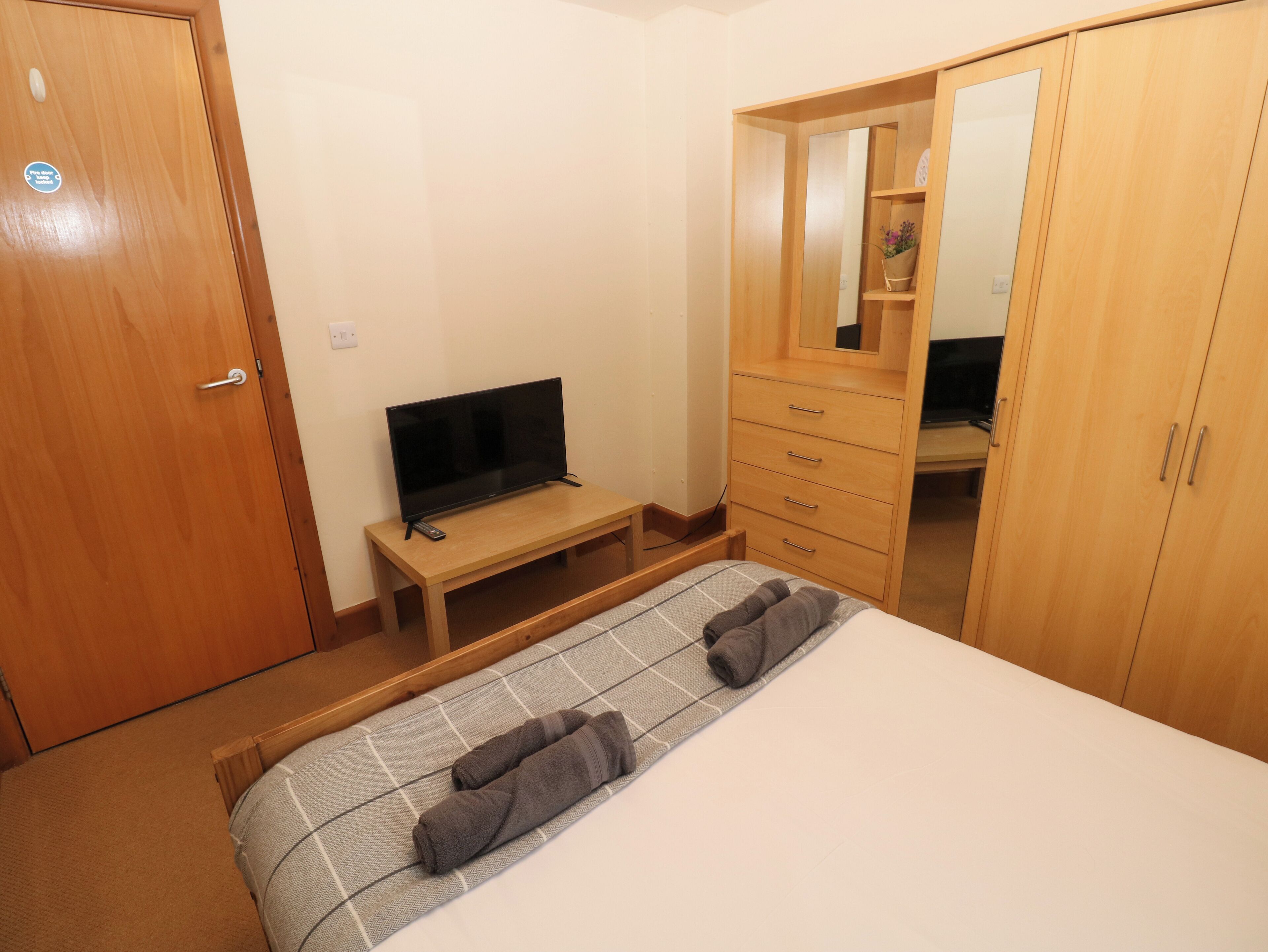 A bedroom with a bed, a TV on a wooden stand, a wardrobe, and a mirror.