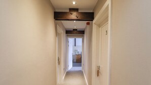A narrow hallway with white walls and a beige carpet. There are two doors on the right side, one of which is open, revealing a small room with a dresser and a lamp. - The Fullerton (Newcastle-upon-Tyne)