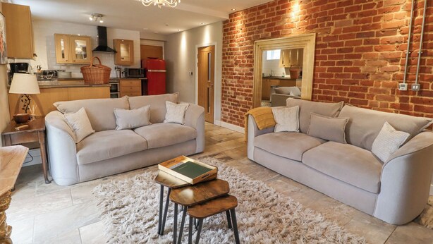 A spacious living room with two sofas, a fluffy rug, and a wooden coffee table. A kitchen with modern appliances is visible in the background. - The Fullerton (Newcastle-upon-Tyne)