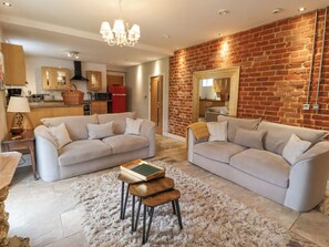 A spacious living room with two sofas, a fluffy rug, and a wooden coffee table. A kitchen with modern appliances is visible in the background. - The Fullerton (Newcastle-upon-Tyne)