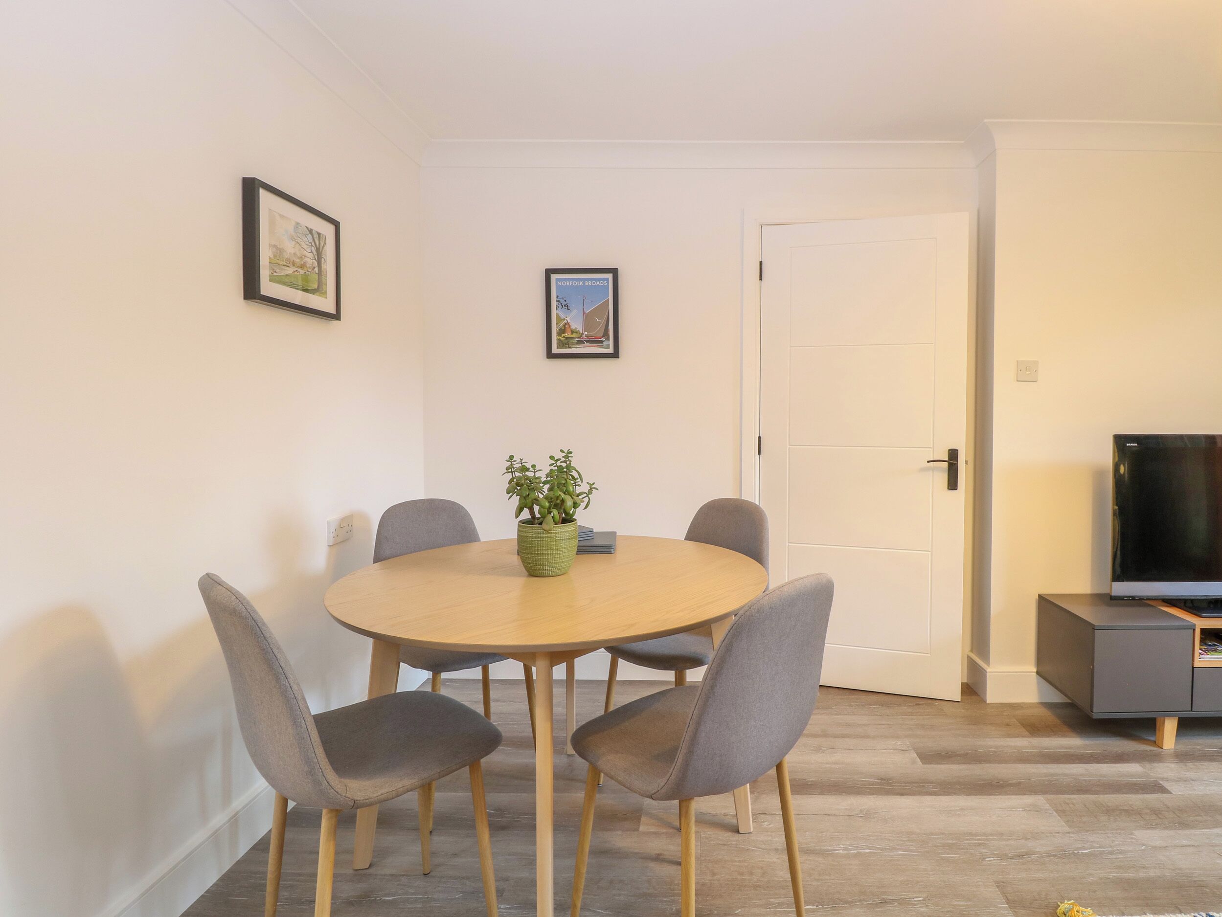 Een eetruimte met een ronde tafel, vier grijze stoelen, een potplant en twee ingelijste foto’s aan de muur.