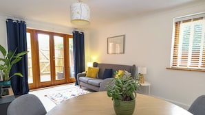 Una sala de estar con un sofá gris, una mesa de comedor de madera, una planta verde y un cojín azul.