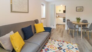 Una sala de estar acogedora con un sofá gris, cojines amarillos y azules, una alfombra colorida, un área de comedor con mesa y sillas, y una cocina al fondo.