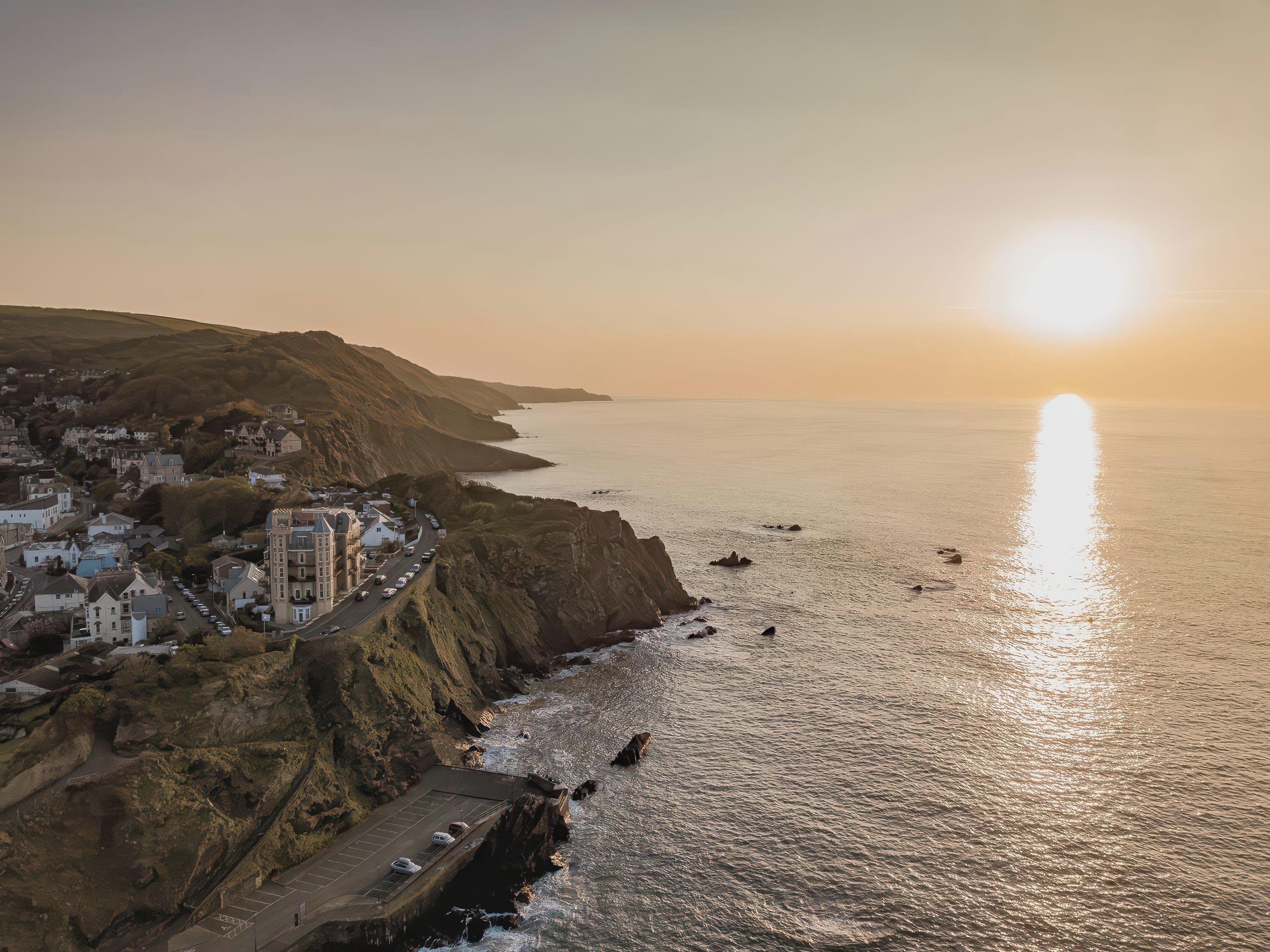 A coastal town with a prominent cliffside, a road leading to a parking area, and a view of the sea at sunset.