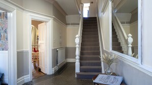 Una amplia escalera con alfombra estampada, un letrero que indica la ubicación y un pasillo con radiador y puerta que da a una habitación con una silla.