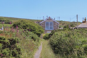 Ein Steinhaus mit großem Fenster und Tür, umgeben von üppigem Grün und einem schmalen Weg, der dorthin führt.