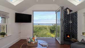 A modern living room with a glass table, a fireplace, and a large window offering a view of greenery and a bridge. - Victory View (Penzance)