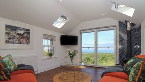 A modern living room with two orange sofas, a glass coffee table, a mounted TV, and a large sliding glass door leading to a scenic view. - Victory View (Penzance)