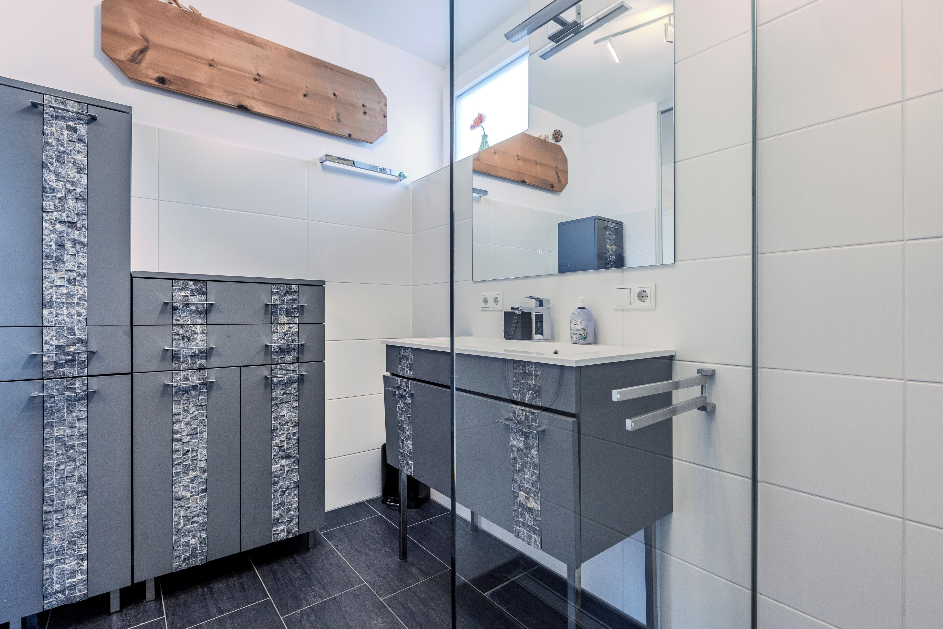 Une salle de bain moderne avec des meubles gris, une douche en verre et une étagère en bois au-dessus du miroir.
