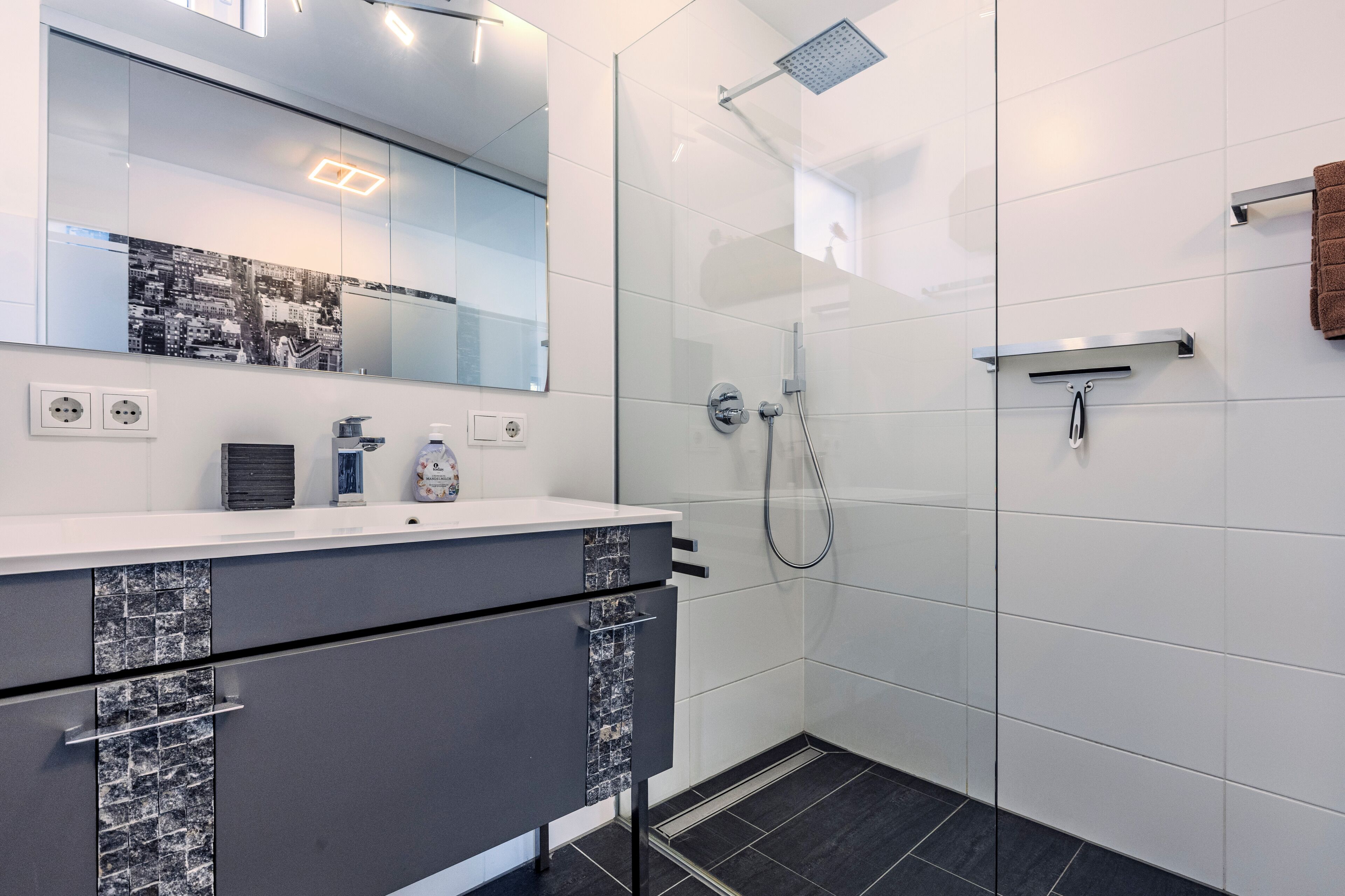 Une salle de bain moderne avec une douche à parois de verre, un mur recouvert de carreaux blancs et un meuble-lavabo gris avec un miroir.