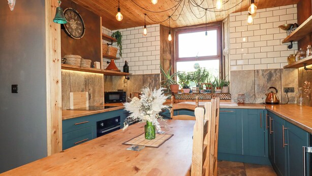 A kitchen with wooden cabinets, a wooden dining table, hanging light bulbs, and a window with plants. - Mermaid's Rest (Newquay)