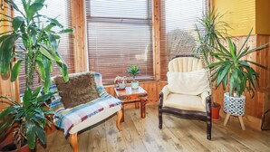A cozy room with wooden flooring, a cushioned chair, a small table with a plant, and large windows with blinds. - Mermaid's Rest (Newquay)