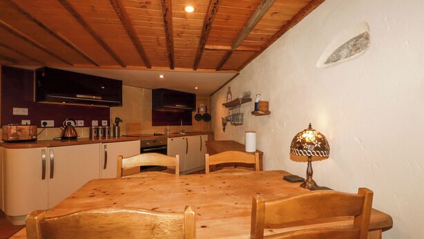 Een houten eettafel met stoelen, een keuken met kasten en apparatuur, en een wandplank met spullen.